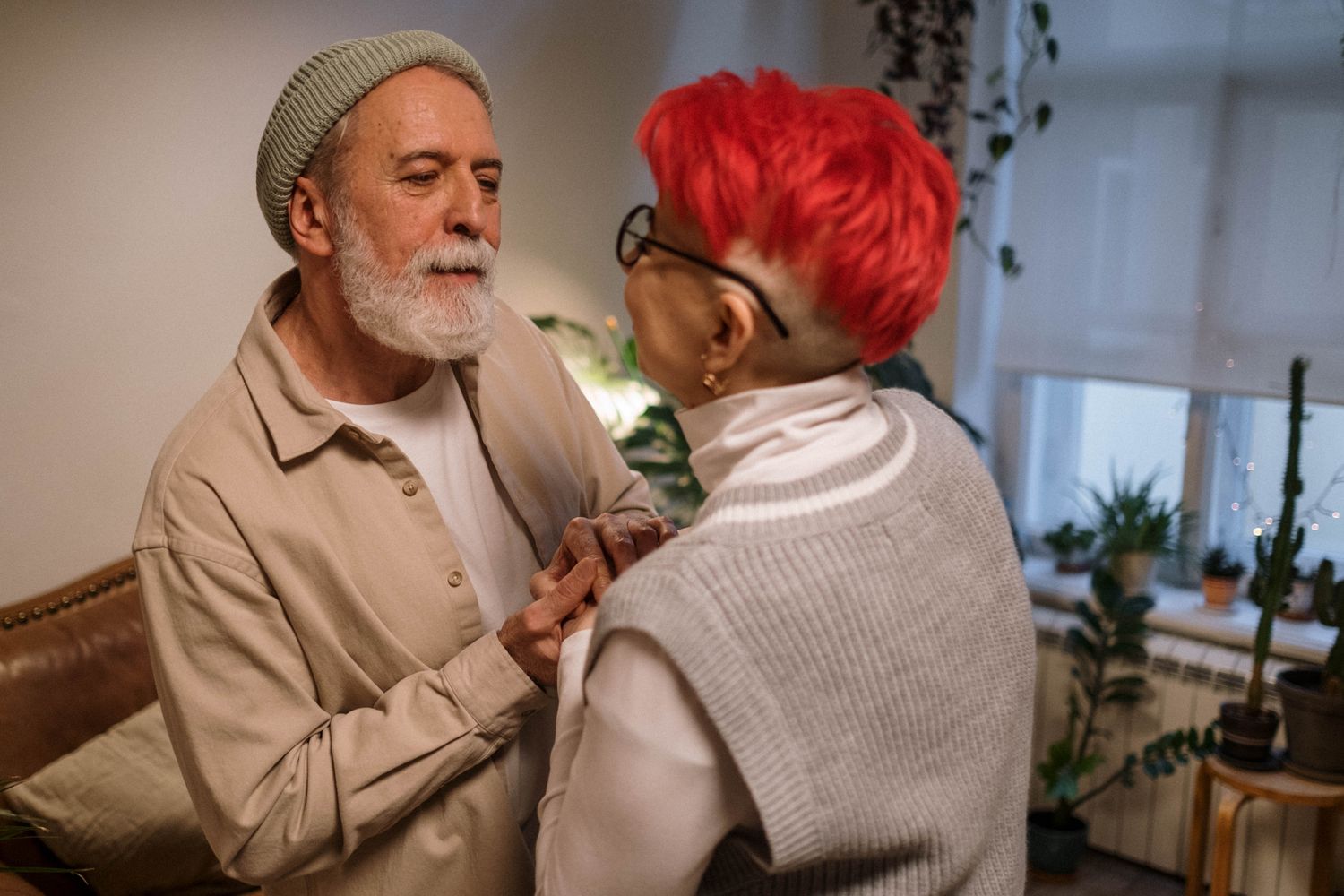 An older husband in a knit cap holds his wife close in their living room